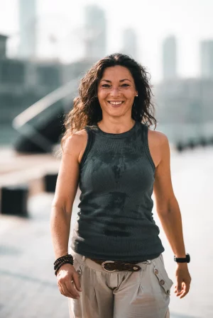 Eine lächelnde Frau mit langen Haaren geht an einem sonnigen Tag draußen spazieren. Sie trägt ein graues ärmelloses Oberteil und eine helle Hose, im Hintergrund sind Stadtgebäude zu sehen.